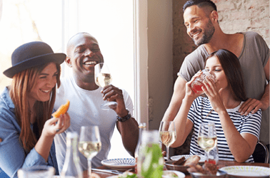Gathering of four diverse laughing adults enjoying wine and appetizers at lunch with each other at restaurant table beside large bright window